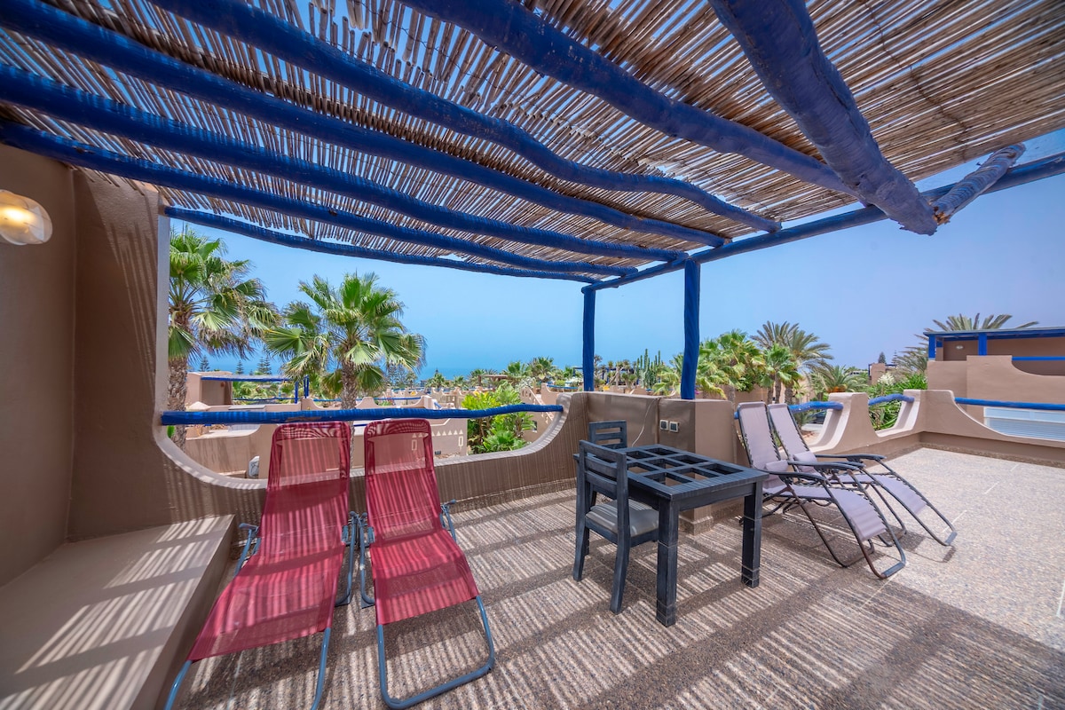 A rooftop terrace is featured, showcasing a shaded seating area with two red lounge chairs and a black table surrounded by palm trees. The expansive view highlights the ocean beyond, with a clear blue sky enhancing the tranquil setting.