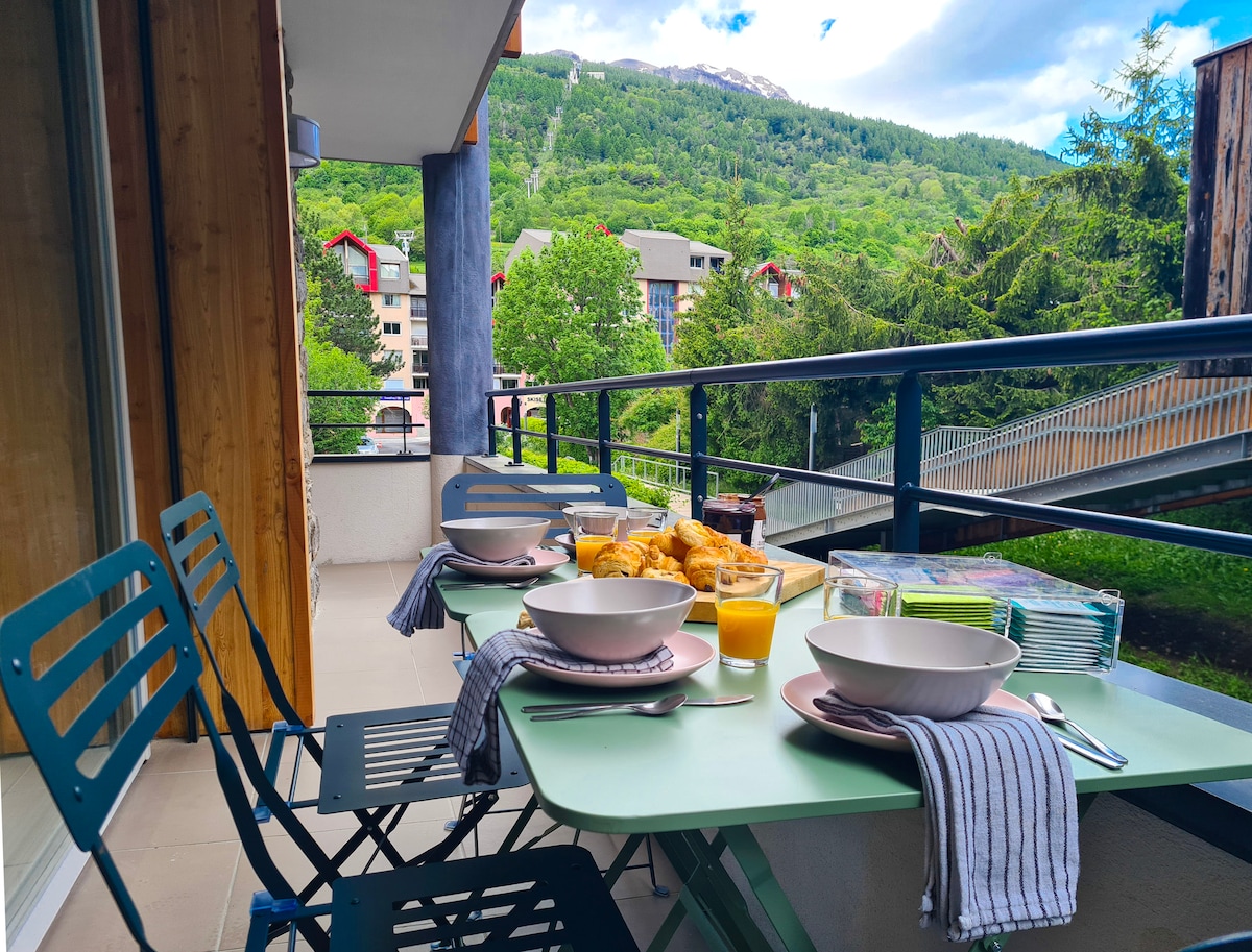 An outdoor terrace is equipped with a green table and four metal chairs, set for breakfast with bowls, juice, and pastries. Lush greenery and mountain views are visible in the background, offering a serene and inviting dining space.
