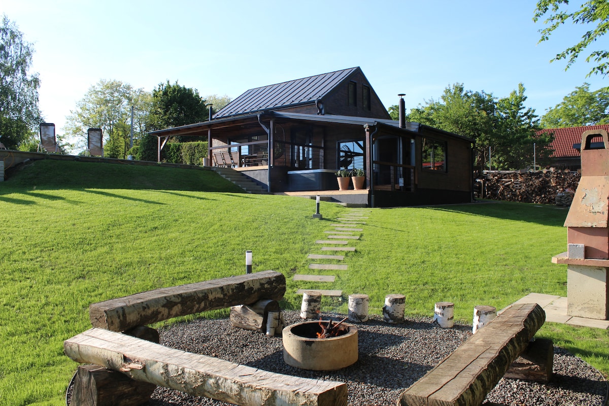 A spacious outdoor area features a well-maintained lawn leading to a modern villa with a covered terrace. Seating is provided around a circular fire pit, and details such as a stone grill and wooden benches offer inviting spaces for gatherings while greenery surrounds the property.