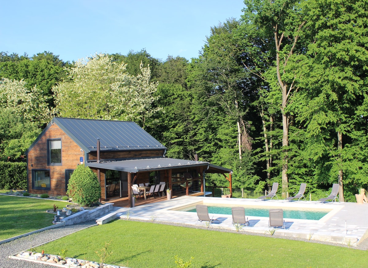 The villa's exterior features a wooden design surrounded by lush greenery. A spacious terrace provides shade and overlooks a seasonal outdoor pool. Several lounge chairs are positioned along the poolside, creating a relaxing atmosphere for outdoor enjoyment.