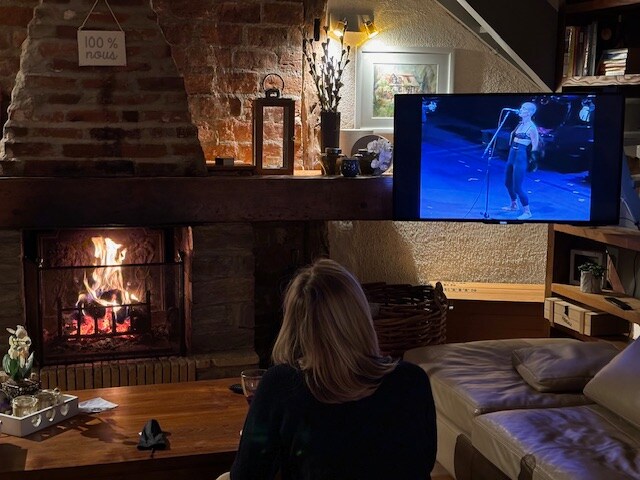 A living room scene is depicted with a warm fire burning in a rustic fireplace. A wooden coffee table, adorned with decorative items, is positioned in front of a comfortable couch. A person is seen from the back, watching a performance on a flat-screen television.