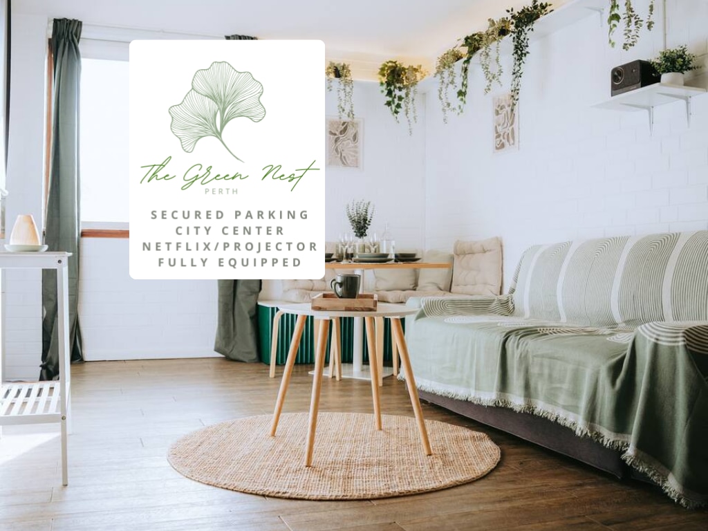 A cozy living room is presented with a light-colored sofa adorned with green and white accents. A small wooden table is at the center, surrounded by leafy plants, enhancing the relaxed atmosphere. A projector setup is visible, ready for entertainment.