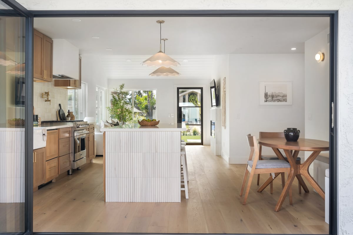 An open-concept kitchen and dining area is visible, featuring a central island with a textured design and modern cabinetry. A wooden dining table with four chairs is situated nearby, complemented by a warm pendant light. Natural light pours in through the large sliding doors, enhancing the airy atmosphere.