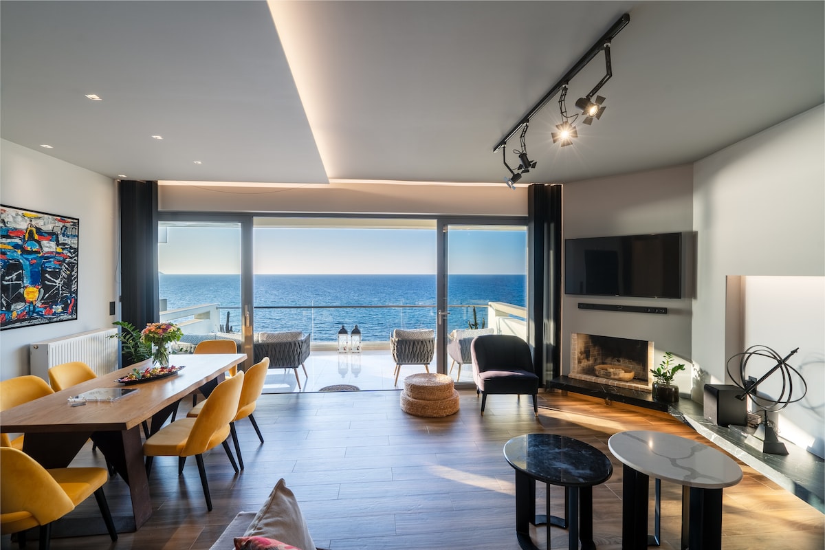 A spacious living area features large sliding glass doors that open to a panoramic sea view. Bright yellow chairs surround a wooden dining table, while a sleek black coffee table and side tables are positioned nearby. A modern fireplace is integrated into the wall, enhancing the contemporary design.