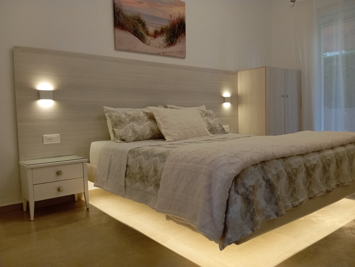 A spacious bedroom is illuminated by soft, warm lighting highlighting a bed with a textured white and floral patterned blanket. A minimalist nightstand is positioned beside the bed, and light-colored cabinets are visible in the background. Curtains are drawn aside, allowing natural light to filter in.