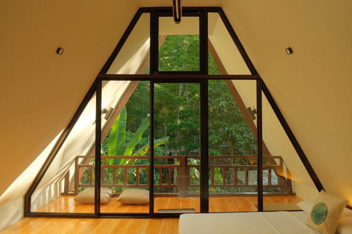 A spacious loft area is featured, highlighted by large glass windows allowing natural light to fill the room. Lush green foliage can be seen just outside, while soft cushions are arranged on the wooden balcony. A minimalist bed rests against the angled wall.