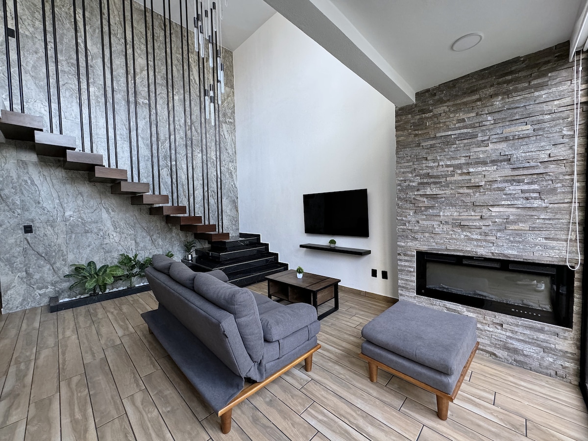 A modern living area features a sleek design with a large flat-screen TV mounted on the wall. Natural light floods the space, highlighting the textured stone fireplace and the staircase with minimalistic railings. A comfortable sofa and ottoman complement the wooden coffee table.