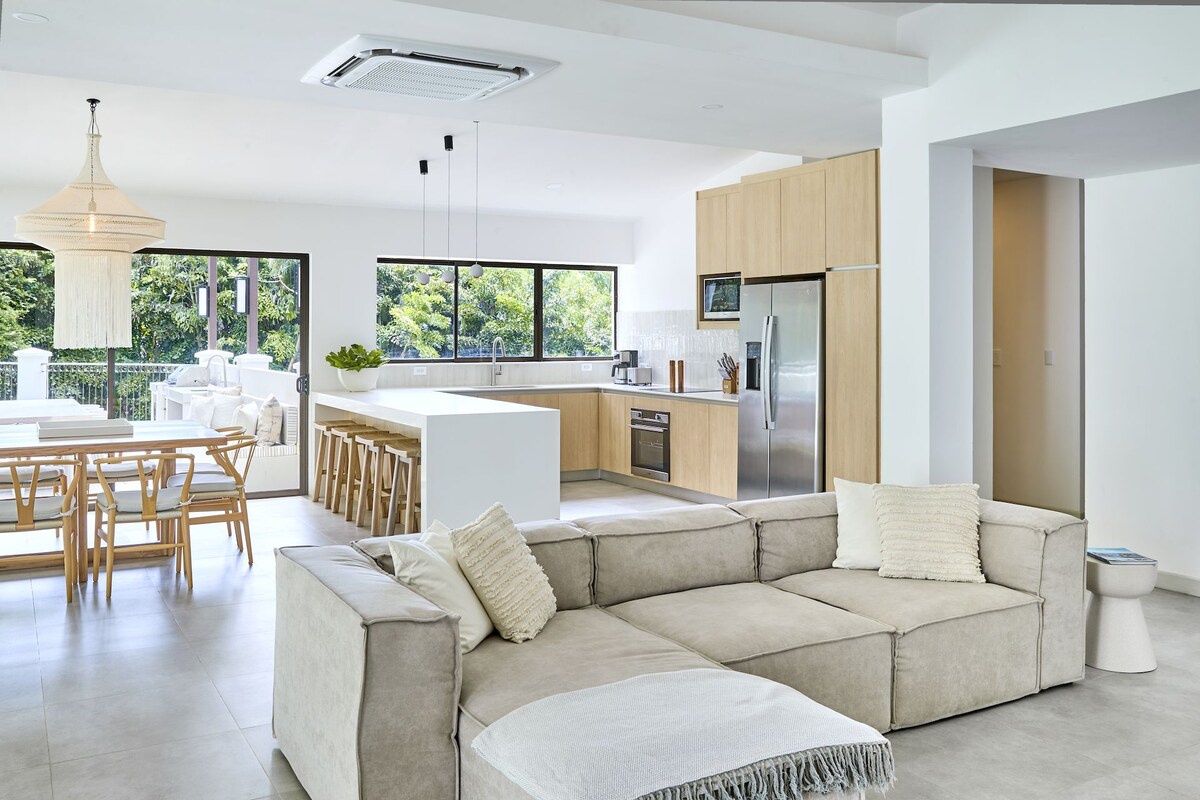 An open-concept living area features a spacious beige sectional sofa, positioned for relaxation. A dining area with wooden chairs and a large table is visible, along with a modern kitchen, stainless steel appliances, and light wood cabinetry. Expansive windows provide views of the lush surroundings.