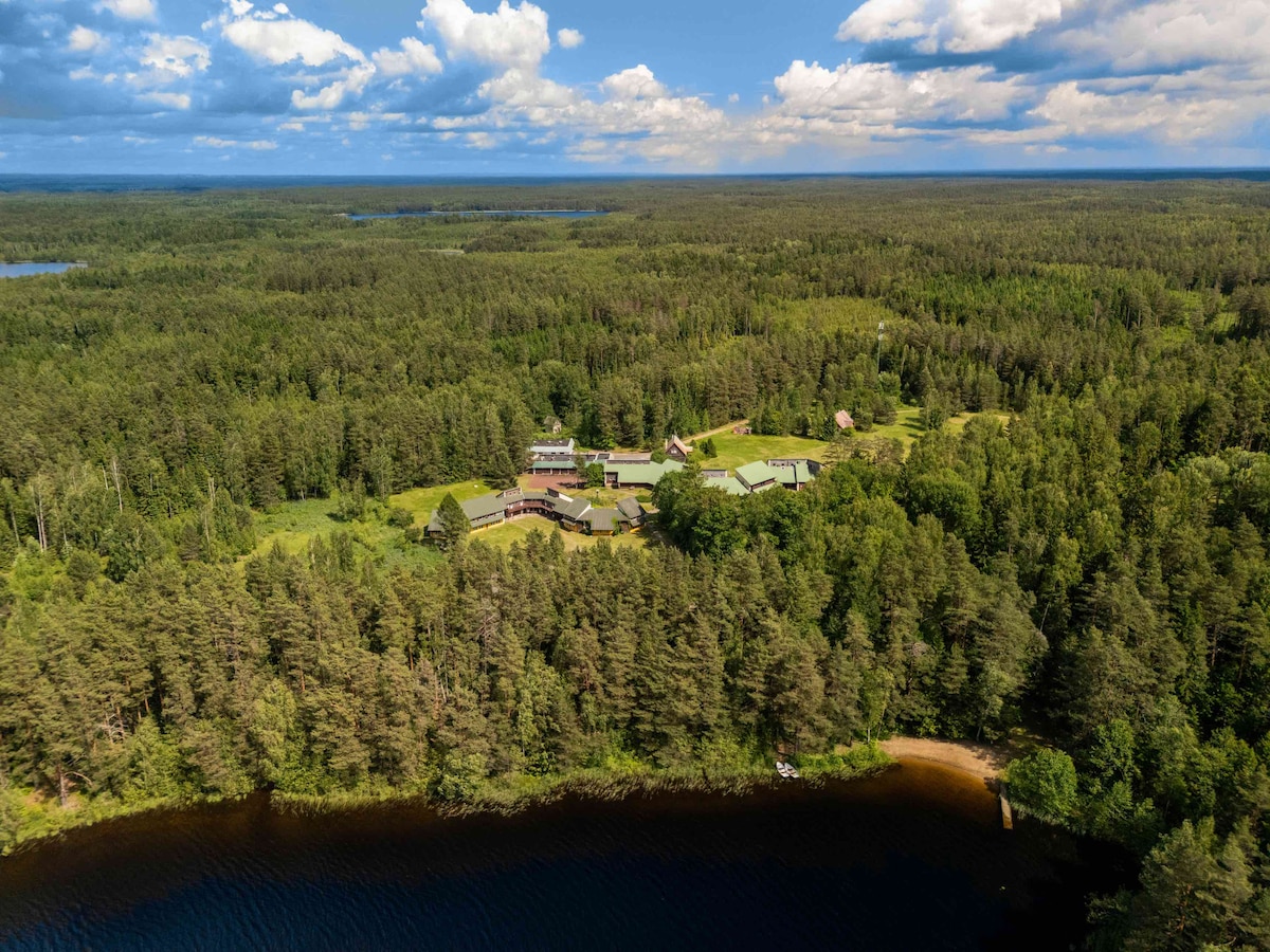 Aerial view captures a serene landscape surrounded by lush green forests and shimmering lakes. A collection of buildings is nestled harmoniously within the natural setting, with a winding shoreline visible along the bottom edge of the image.