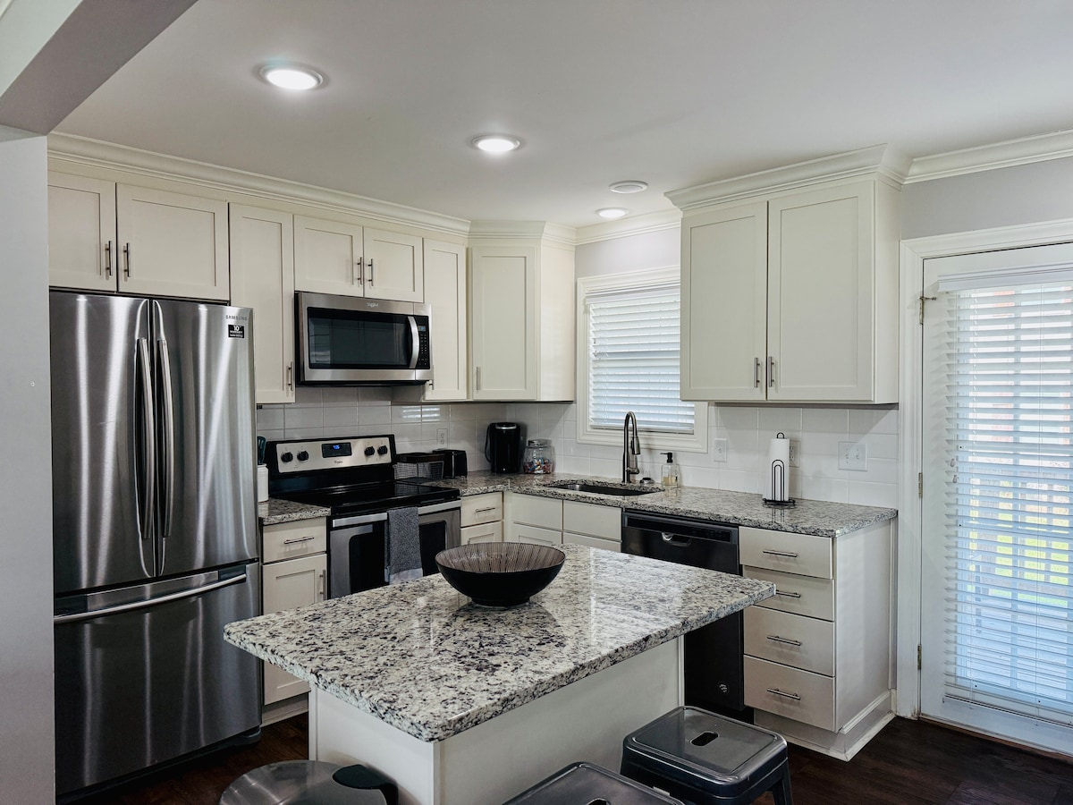 The kitchen features modern stainless steel appliances, including a refrigerator and microwave. Light-colored cabinetry contrasts with dark countertops, while a central island provides additional workspace and seating. Natural light enters through a nearby window, illuminating the room. Blinds are drawn to offer privacy.