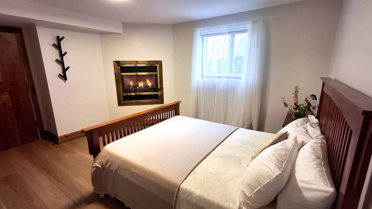 A cozy bedroom features a queen-sized bed with crisp white linens and plush pillows. A gas fireplace is positioned against the wall, providing warmth and ambiance. Natural light enters through a nearby window, complemented by soft, sheer curtains, while delicate flowers add a touch of nature.