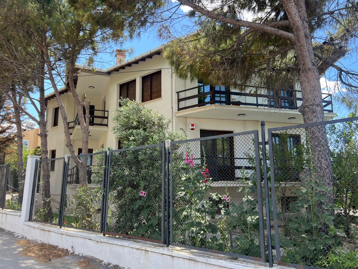 A two-story building is displayed amidst a natural setting, surrounded by tall trees. A gated area with a fence frames blooming flowers and greenery, while balconies overlook the outdoor space, offering a welcoming appearance.