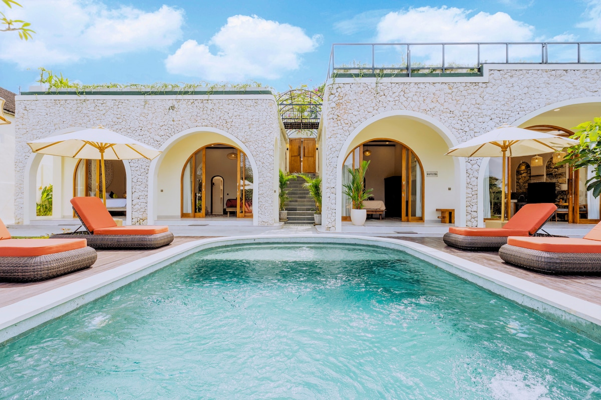 A private swimming pool is framed by elegantly arched structures. Sun loungers with cushions are positioned nearby, inviting relaxation. Lush greenery and a rooftop terrace are visible, complementing the serene atmosphere of the villa surrounded by natural beauty.