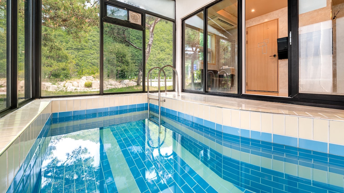 An indoor pool with clear blue tiles reflects natural light from large glass windows. Lush greenery and outdoor seating are visible through the windows, providing a serene view. The pool area is designed for relaxation and enjoyment, inviting guests to swim at their leisure.