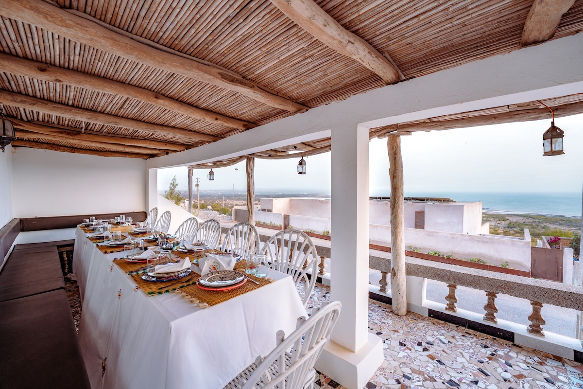 An open-air dining area is presented with a long table, set elegantly for a meal. Surrounding the space are white chairs and a low seating area, with coastal views visible through large windows. The ceiling features wooden beams, adding a natural touch.