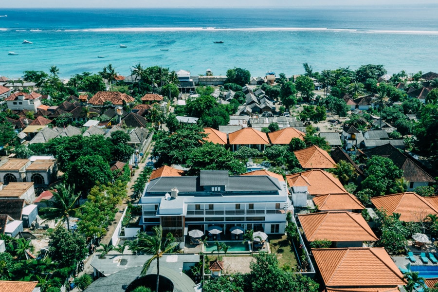An aerial view captures the layout of the resort within a lush environment. The arrangement of terracotta-roofed buildings is visible amidst greenery, with the shimmering ocean in the background. Shared pools and nearby beach access are indicated by the surrounding structures and foliage.