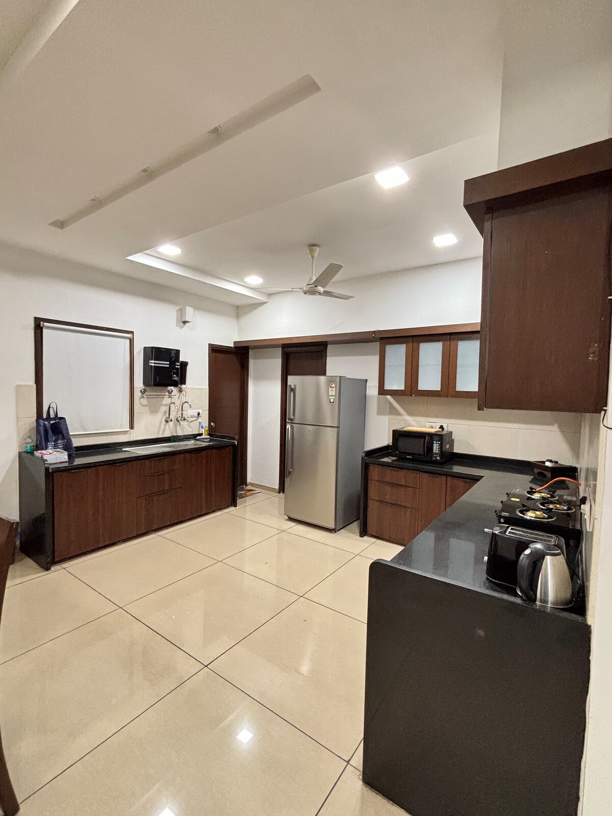 A modern kitchen is depicted with dark wooden cabinets and sleek black countertops. Stainless steel appliances include a refrigerator and microwave. The space is well-lit with ceiling lights, and a countertop coffee maker is positioned near the edge. A window allows natural light to enter.