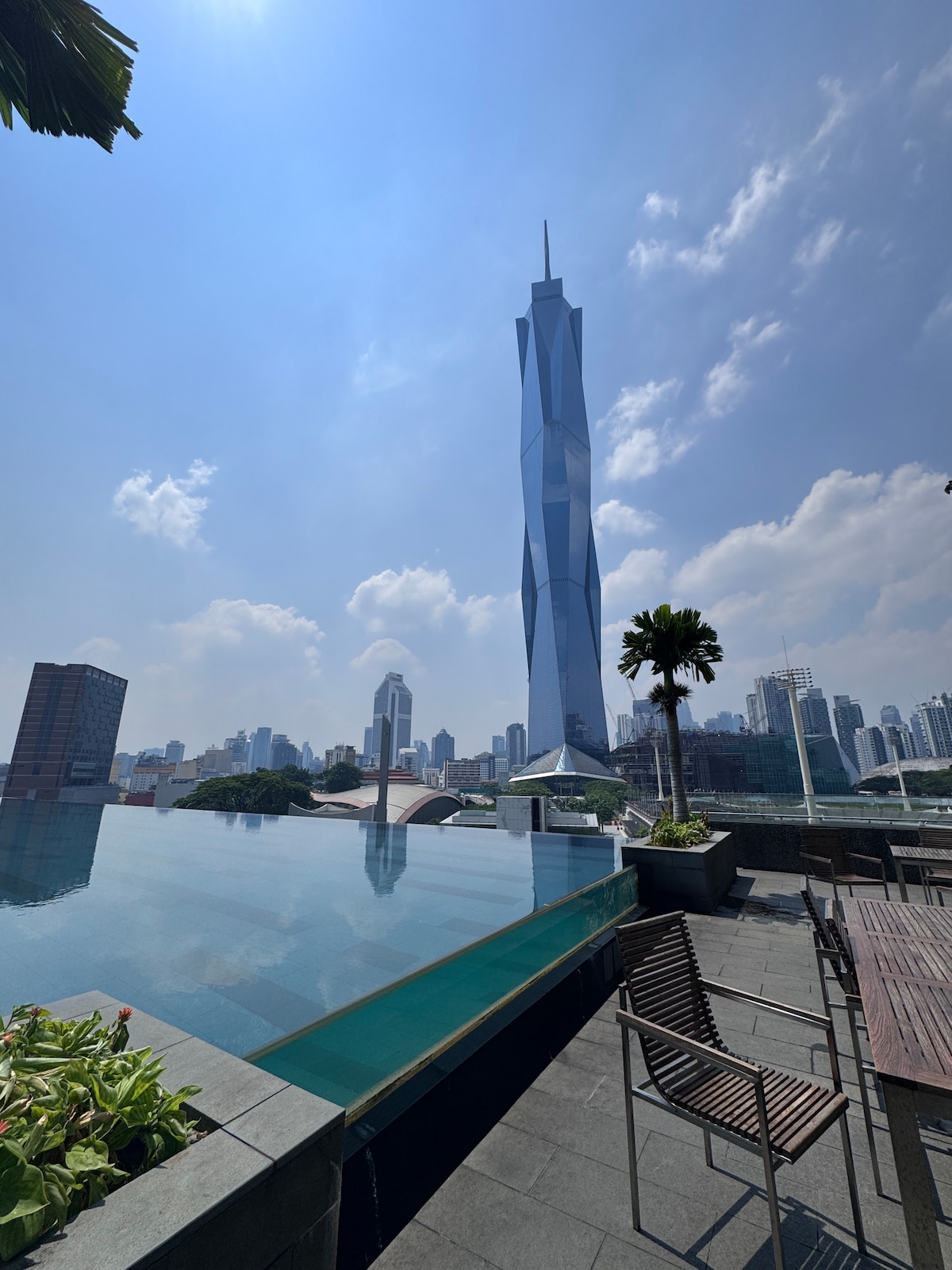 A serene rooftop pool reflects the sky, with the striking Merdeka 118 tower rising prominently in the background. Sunlight bathes the area, and modern seating arrangements are visible, surrounded by tasteful landscaping that adds greenery to the urban setting.