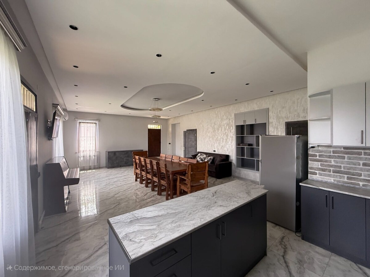 An open-plan living area features a spacious dining table surrounded by wooden chairs, set against a backdrop of light-colored walls. A modern kitchen with sleek cabinets is visible, along with a cozy seating area that includes a dark couch. Natural light illuminates the space through large windows.