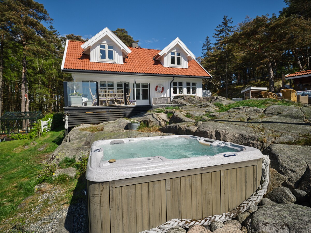 A hot tub is positioned on a wooden deck, surrounded by natural stone and green grass. In the background, a charming two-story house with a red roof and large windows offers views of the scenic outdoor space. Tall trees frame the property, contributing to a serene atmosphere.