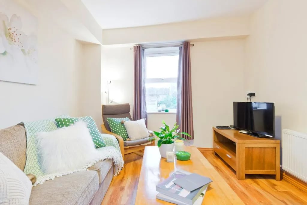 A welcoming living area features comfortable seating, including a sofa adorned with cushions and a cozy throw. Natural light enters through a window, complemented by subtle curtains. A wooden coffee table holds magazines, while a green plant adds a touch of nature to the space.