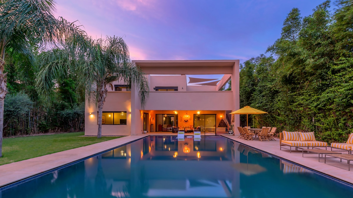 The modern villa is set against a vibrant sunset, featuring large windows that reflect the colors of the sky. A sparkling pool stretches along one side, bordered by palm trees and outdoor seating areas with bright striped cushions and a shaded dining space under a yellow umbrella.