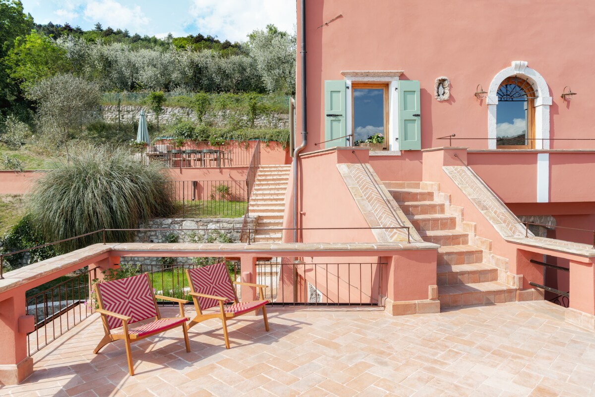 An outdoor terrace features two striped lounge chairs positioned for relaxation, with surrounding vibrant greenery and a staircase leading to upper levels. The warm pink walls of the building provide a contrasting backdrop, while wooden shutters frame an inviting doorway.