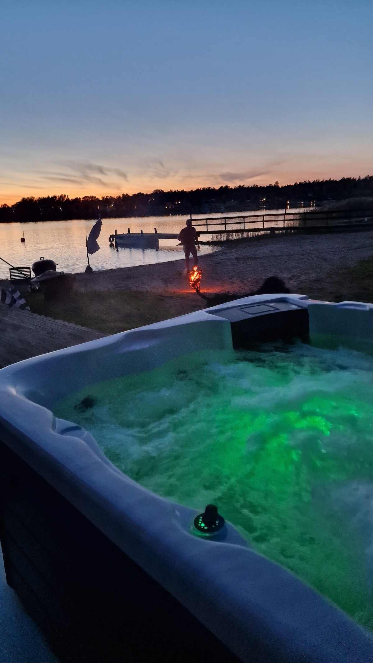A hot tub with illuminated jets is positioned on a deck overlooking a tranquil lake at sunset. Silhouetted against the colorful sky, a person stands near a fire pit on the sandy beach, with a dock visible in the background.