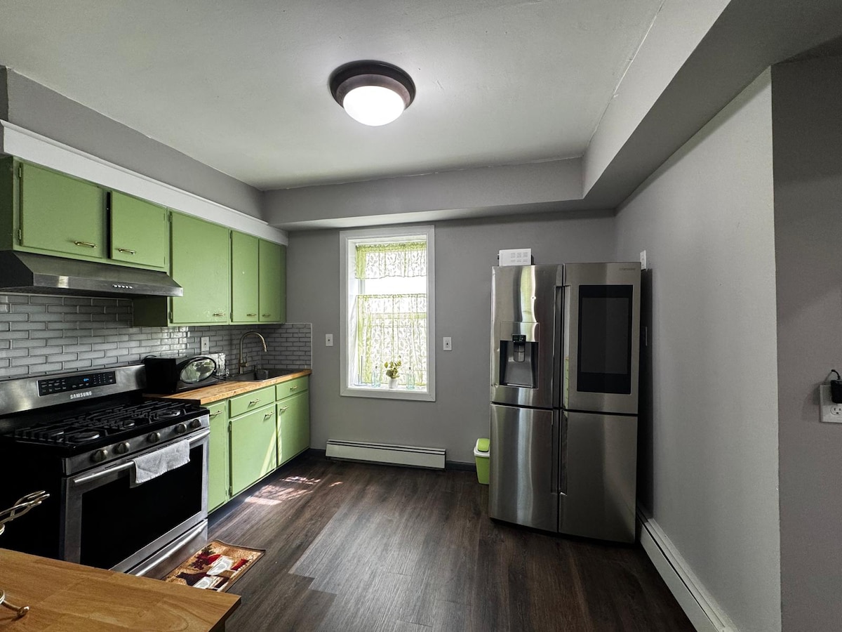A modern kitchen is presented with green cabinetry and a stainless-steel refrigerator. A gas range is located beneath a sleek extractor hood. Natural light filters through a window, illuminating the wooden countertop and tile backsplash. The flooring features dark laminate, adding to the contemporary feel.