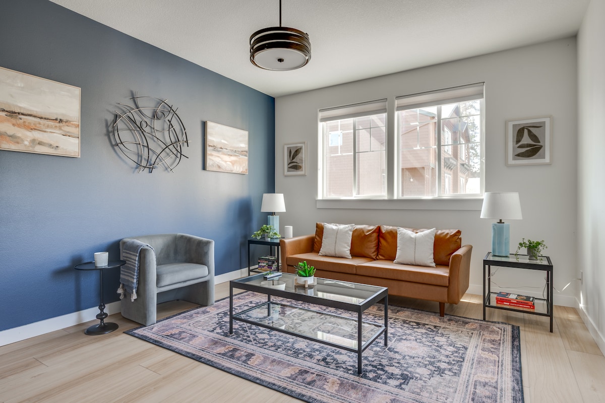 A comfortable living area is featured, showcasing a brown sofa adorned with accent pillows. Two lamps illuminate the space, while a coffee table sits atop a patterned rug. A blue accent chair and wall art complement the blue-painted wall, with ample natural light from the large windows.