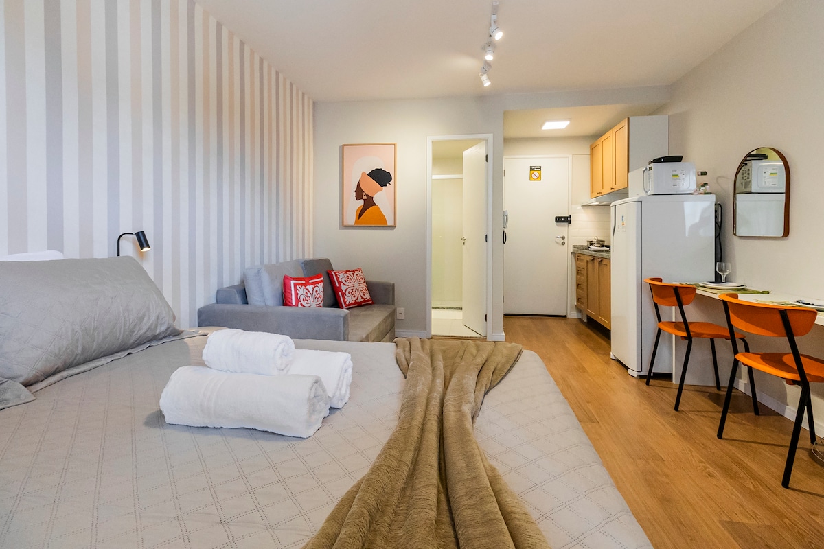 A well-appointed studio apartment features a spacious bed covered with soft linens. A seating area with a gray sofa and decorative pillows is visible. The kitchenette is equipped with essential appliances, while a minimalist artwork adorns the wall, adding a touch of color to the neutral tones.