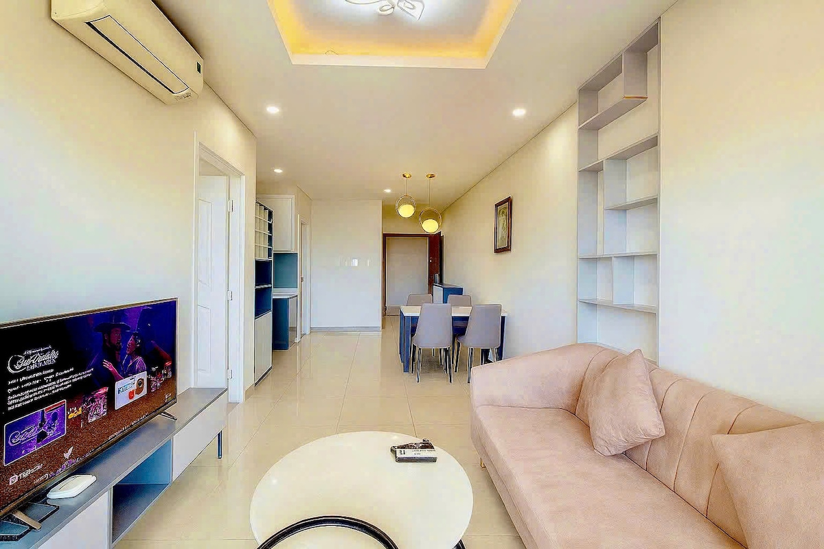 A modern living area features a soft beige sofa, complemented by a circular coffee table. A large television is visible, and a dining area with blue chairs is situated in the background. Natural light illuminates the space through well-placed fixtures.
