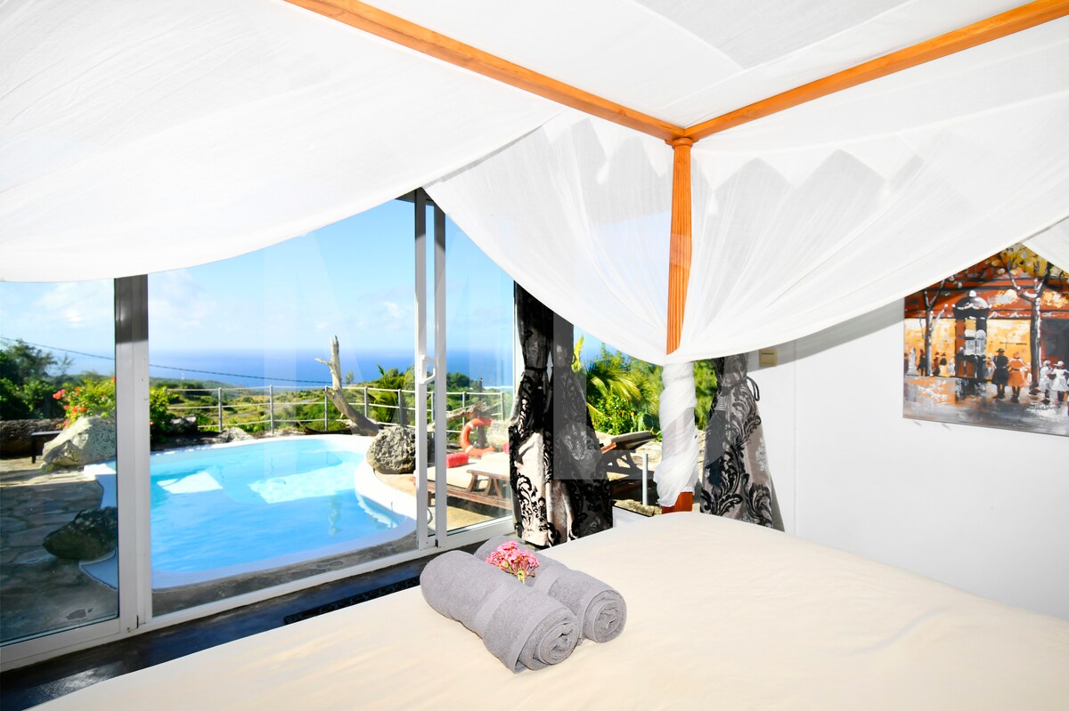 A spacious bedroom features a four-poster bed with flowing drapes. Large glass doors offer views of a private pool and the surrounding landscape. Fresh towels are neatly arranged at the foot of the bed, with vibrant flowers providing a touch of color.