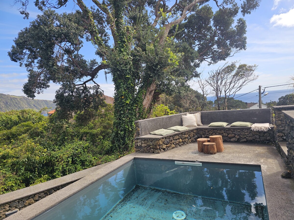 An inviting outdoor space features a small swimming pool framed by natural stone. Near the pool, cushioned seating along a stone wall provides a relaxing area, with wooden stools placed beside it. Lush greenery and trees complement the serene view of the surrounding landscape.
