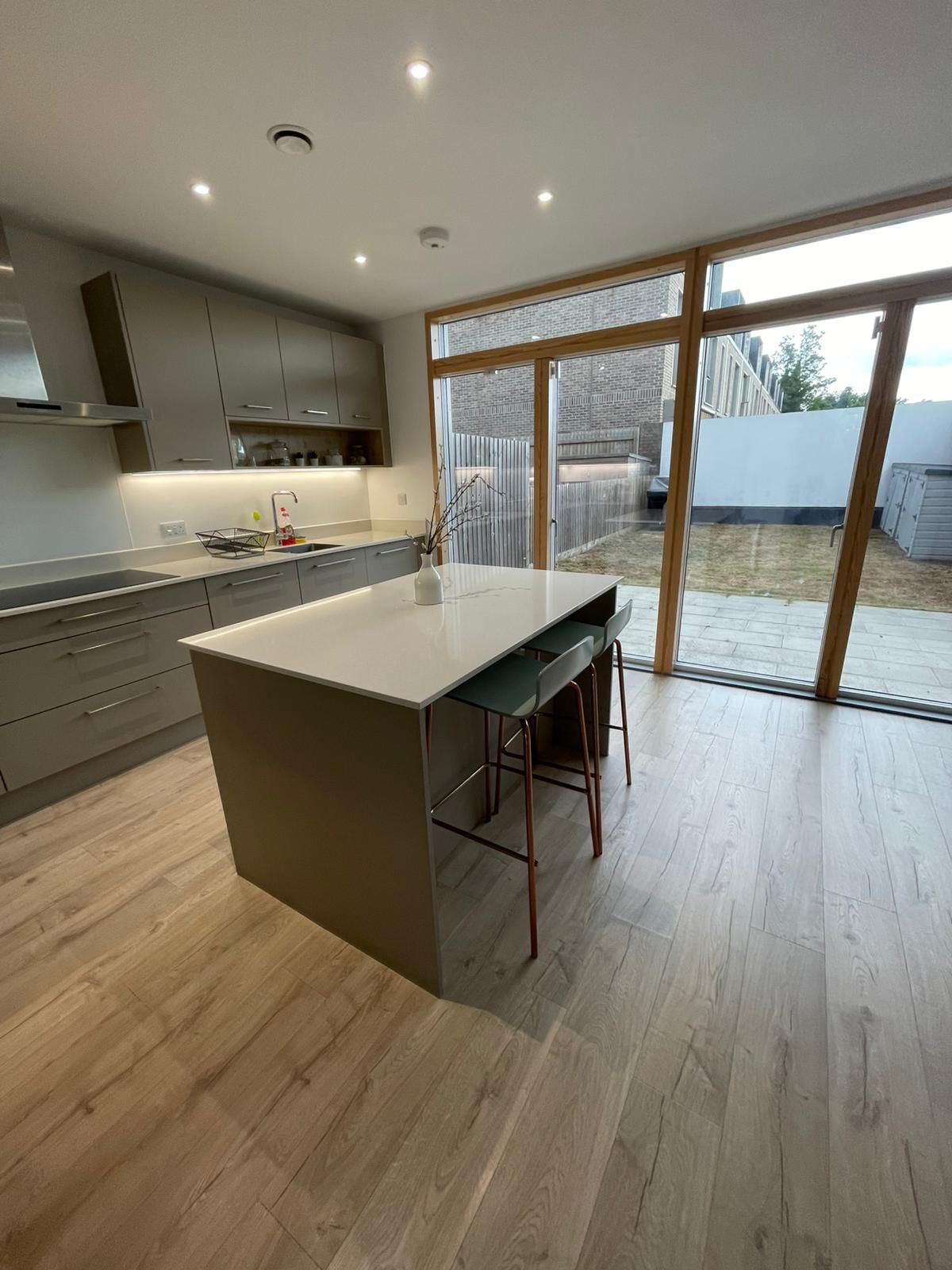 A modern kitchen is presented with sleek cabinetry and a central island. Large windows allow natural light to fill the space, highlighting a light wood floor. Bar stools are positioned neatly at the island, offering a casual dining option. An outdoor area is visible through the glass doors.
