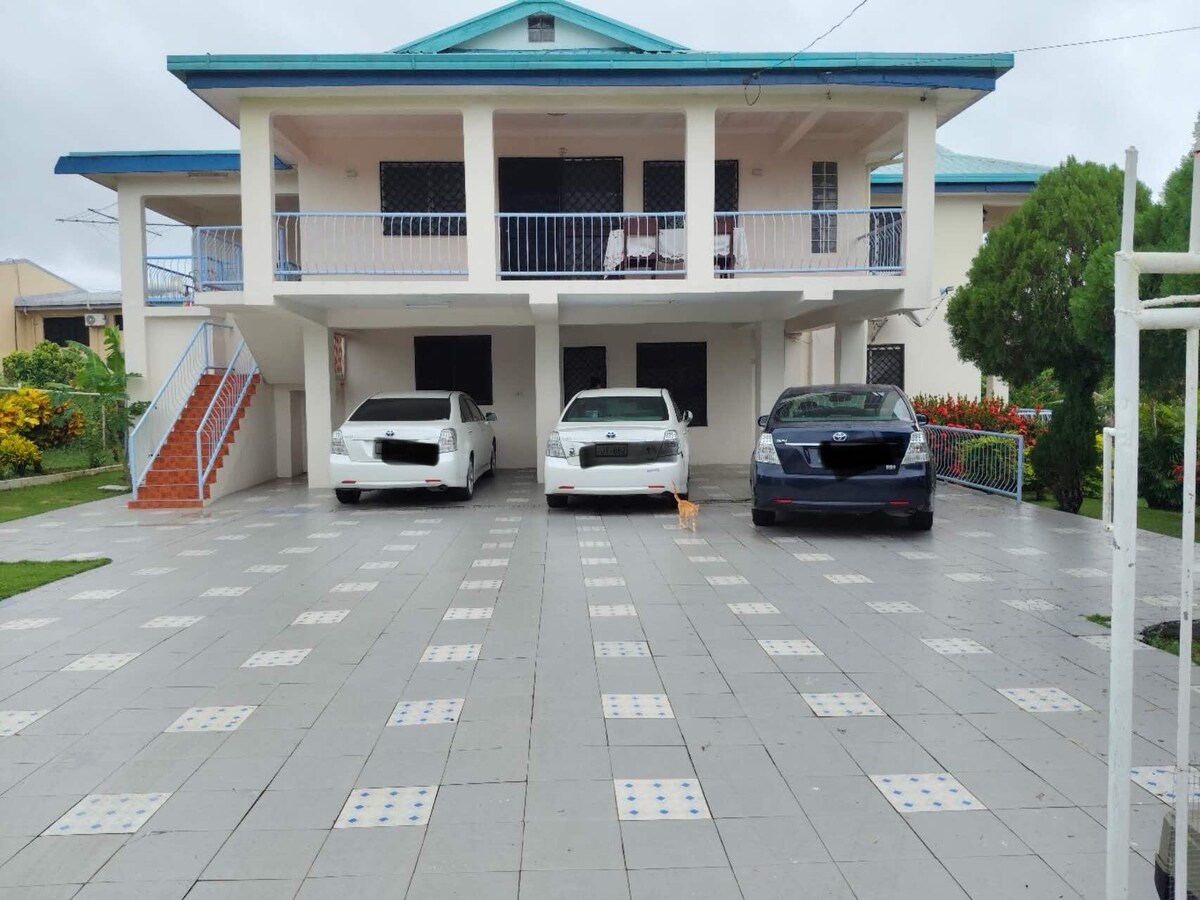 A spacious parking area is shown in front of a two-story building, featuring three parked cars. The area is paved with square tiles, bordered by colorful gardens and greenery, enhancing accessibility. The structure's balcony is visible, offering outdoor seating options.