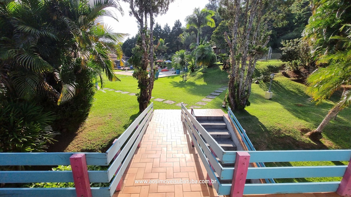 A view from a pathway highlighted by colorful railing, leading towards a manicured lawn. Lush greenery and palm trees frame the scene, guiding the eye towards a swimming pool in the background, providing an inviting outdoor space.