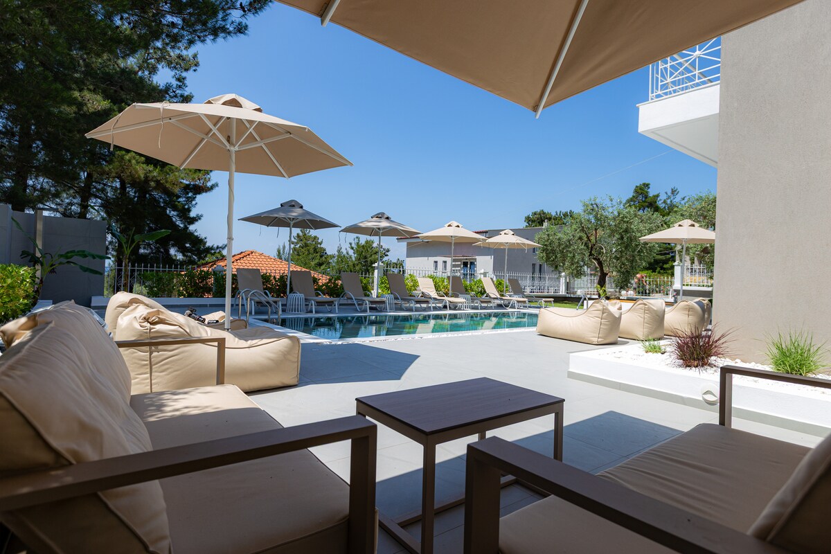 A spacious outdoor terrace features comfortable lounge seating, with a couple of umbrellas providing shade. The inviting pool is lined with sun loungers, surrounded by greenery, while olive trees and pines can be seen in the background.
