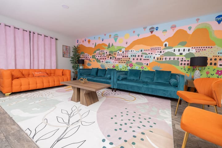 Светла и шарена дневна соба со смели портокалови и темни каучи, живописен мурал на живописно село со балони со топол воздух и модерен декор. Совршен, весел простор за опуштање или собирање со пријателите за време на вашиот престој!