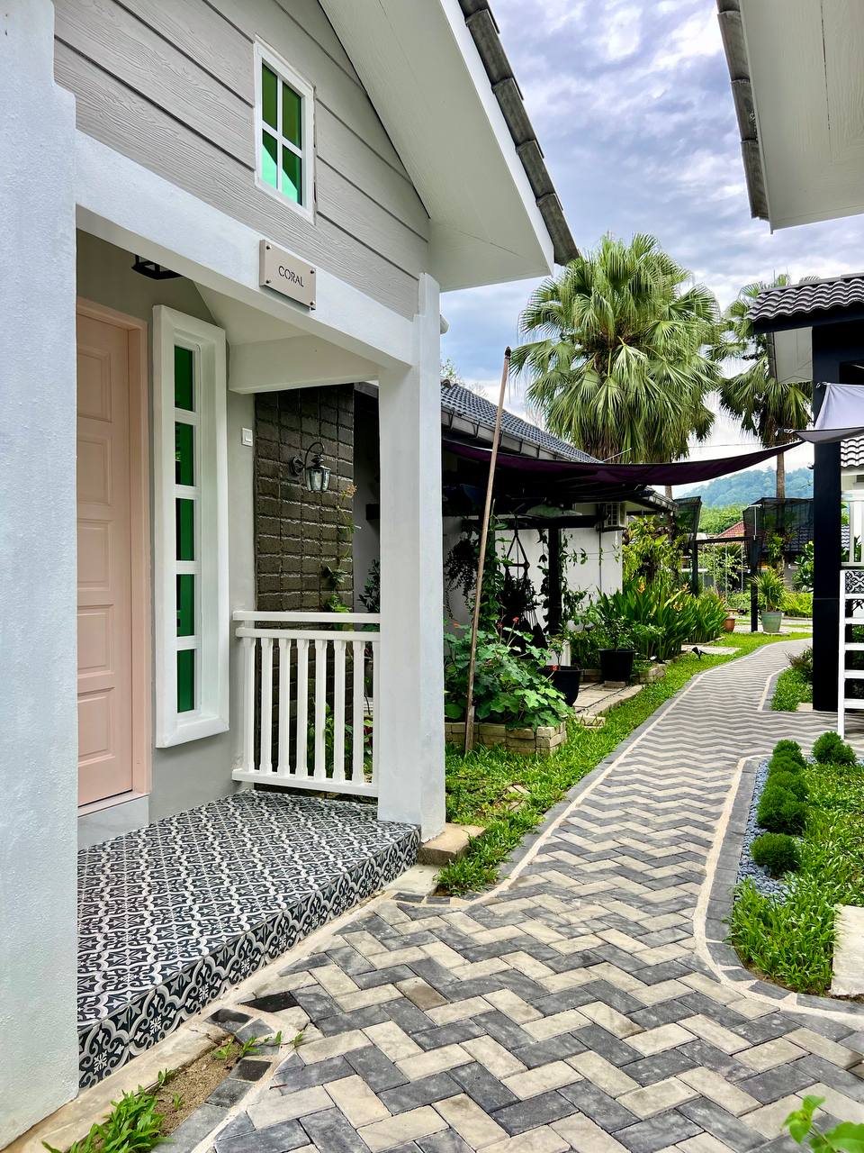 A charming pathway leads to the entrance of a white house with pastel accents. Lush greenery borders the path, creating a serene environment. The house features a decorative porch with a patterned tile floor and large windows that complement the tranquil setting.