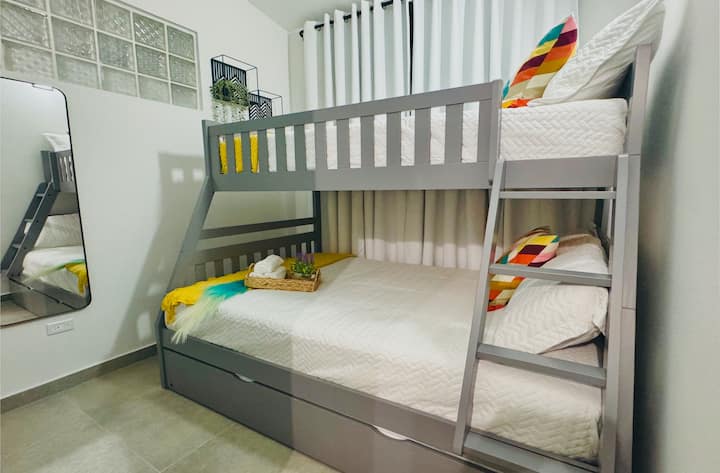 Ova spavaća soba ima sivi krevet na kat s bijelom posteljinom, žute i šarene geometrijske detalje te ugrađene ladice za pohranu. U sobi se nalazi stalak za odjeću, klima-uređaj i bijele zavjese s modernim ukrasima.