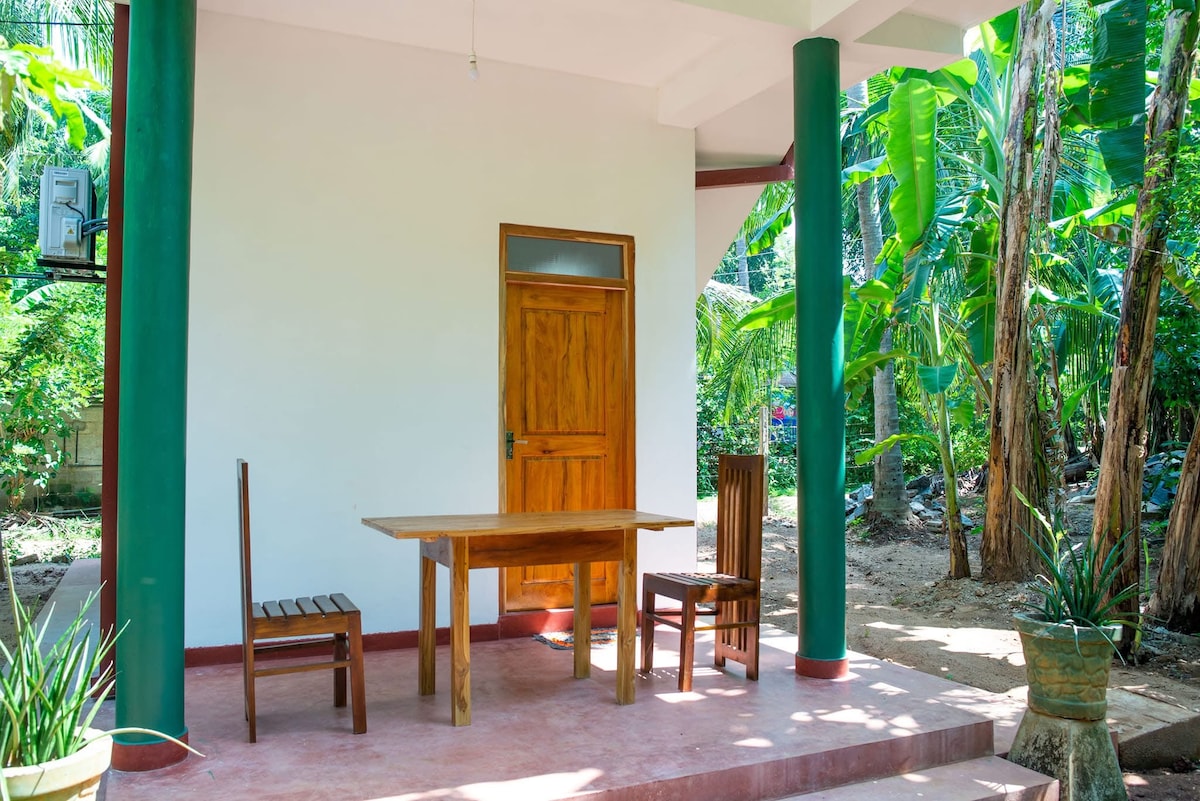 A shaded outdoor seating area features a wooden table surrounded by two wooden chairs. Lush greenery from nearby plants and trees provides a natural backdrop, while the space is complemented by red and green accents in the structure.
