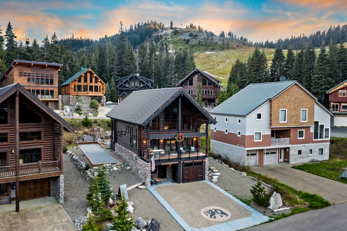 The Black Bear Lodge showcases a spacious, modern cabin with a rustic wooden facade. Surrounded by towering trees and warm sunset hues, the property features unique architectural details, a paved driveway, and nearby access to additional cabins, creating a serene mountain retreat ambiance.