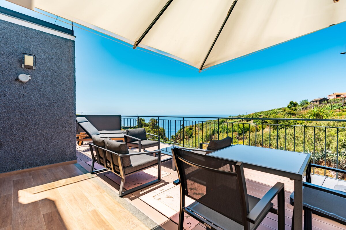 An outdoor terrace features a stylish table and chairs, complemented by sun loungers. An umbrella provides shade, while picturesque views of the ocean and surrounding hills are presented in the background, showcasing a clear blue sky.