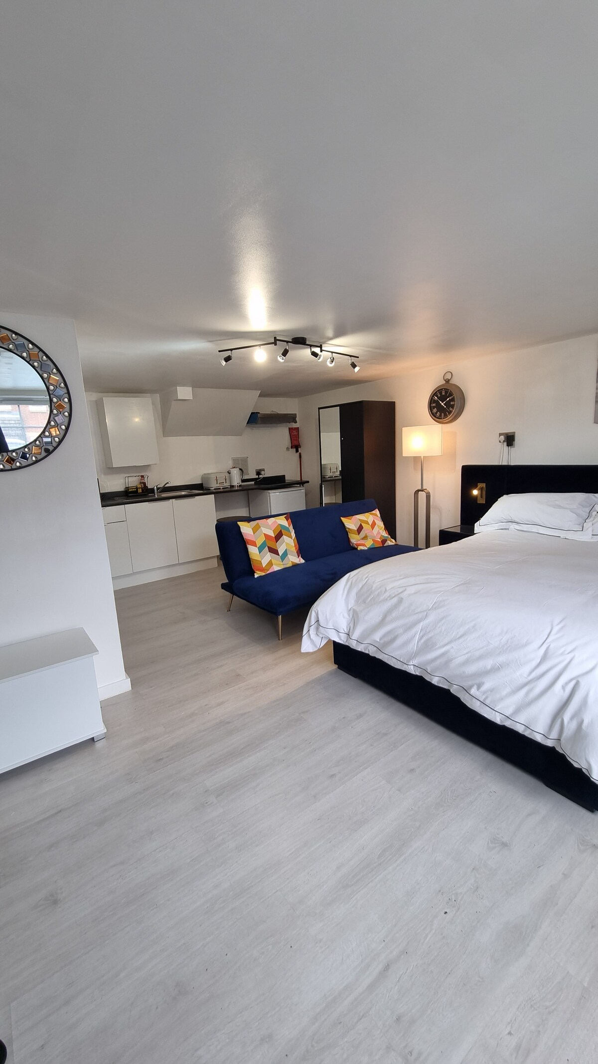 A spacious studio flat is highlighted by a comfortable bed and a sofa, both adorned with decorative cushions. A well-equipped kitchen area is visible in the background, while modern fixtures and lighting contribute to the clean, bright environment.