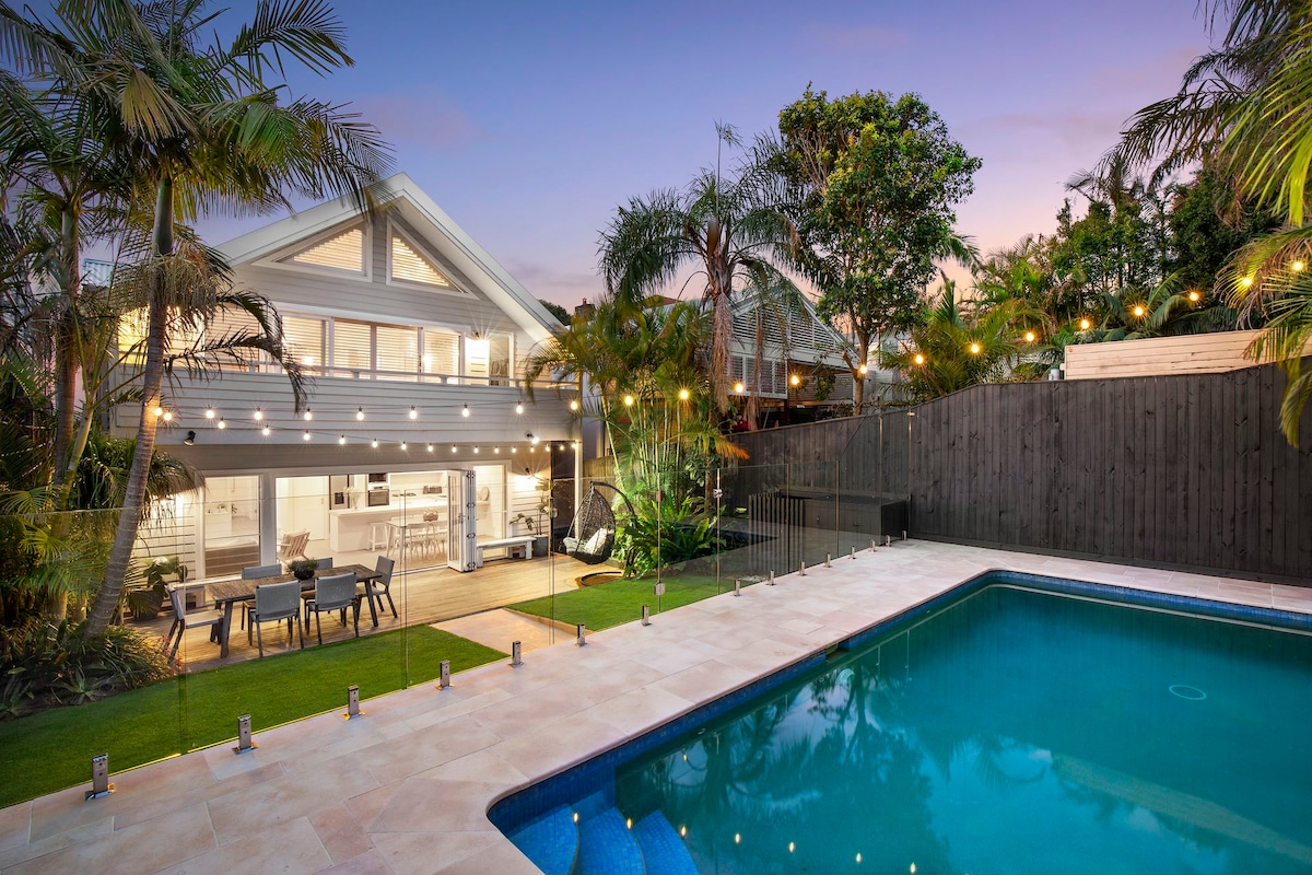 A spacious outdoor area features a clear swimming pool surrounded by light-colored stone paving. Lush greenery and palm trees enhance the tropical atmosphere. The house, with its raked roof, is visible in the background, showcasing large windows that invite natural light.