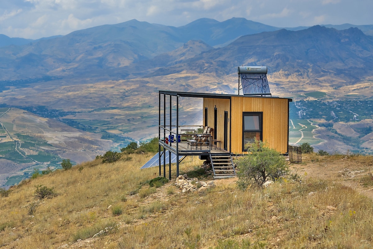 A modern cabin is situated atop a hillside, featuring large windows that offer panoramic views of the surrounding valleys and mountains. A small deck extends from the structure, equipped with outdoor seating, and the natural landscape is marked by golden grasses and scattered shrubs.