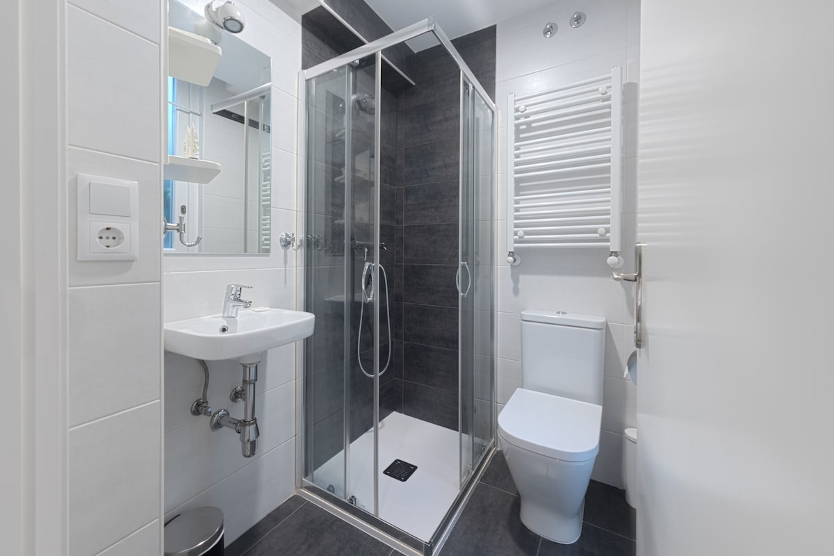 A modern bathroom features a glass-enclosed shower with a sleek design. A white sink and toilet are complemented by a mirror above the sink. Chrome towel racks are installed on the wall, and the flooring is finished in dark tile, creating a contemporary aesthetic.