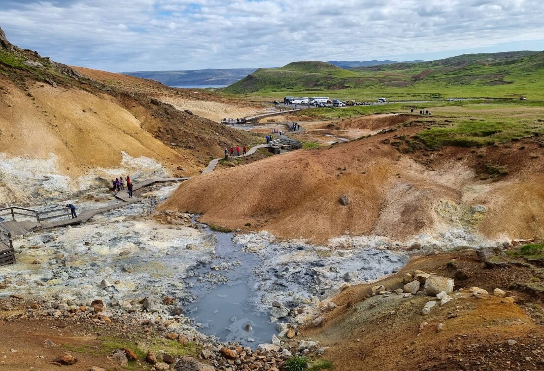 An expansive geothermal area is depicted, featuring colorful mineral-rich soil and steam rising from natural hot springs. Wooden walkways traverse the terrain, with several visitors exploring the landscape. Lush green hills and distant mountains frame the scene, under a partly cloudy sky.