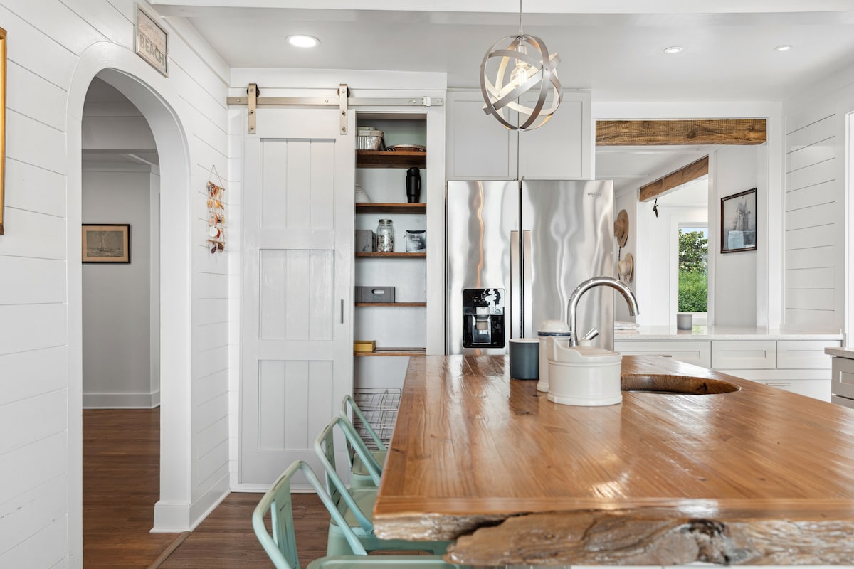 A modern kitchen showcases a large, rustic wooden island, featuring a central sink and inviting seating. Stainless steel appliances are visible, alongside open shelving displaying dishware. Natural light floods the space through windows, enhancing the clean, bright atmosphere of this thoughtfully designed area.